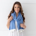 Dreamsoft Organic Cotton Travel Scarf French Blue in color with white band shown as scarf on smiling model in white dress in front of white decorative wall.