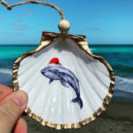Santa's Sea Creatures Whale large seashell ornament with painted whale in santa hat. Ornament is trimmed in gold leaf and being held up with beach in the background.